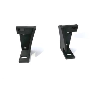 May include: Two black, angular brackets are displayed against a white background. Each bracket has a unique shape with a rectangular base, a curved central section, and a top section with a rectangular cutout. Two circular holes are visible on the curved section.
