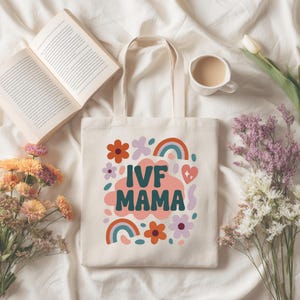 Może przedstawiać: Kremowa torba typu tote z napisem "IVF MAMA" w kolorze morskim i różowym, otoczona kwiatowymi i tęczowymi grafikami. Torba leży na białej powierzchni z otwartą książką, kwiatami i filiżanką kawy.