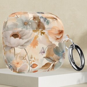 May include: A clear AirPods case with a floral pattern in pastel colours, including white, peach, and blue flowers, and a butterfly. It has a silver ring attachment and is displayed on a white platform.
