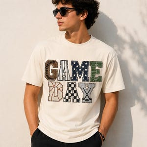 Peut inclure: Un t-shirt à manches courtes de couleur crème avec le texte GAME DAY en grandes lettres de style bloc. Chaque lettre présente un motif différent sur le thème du sport, incluant des éléments de baseball, basket, football et hockey.