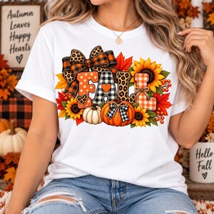 Peut inclure: Un t-shirt blanc avec un motif du mot FALL en lettres à motifs, décoré de tournesols, de feuilles d'érable rouges, d'une citrouille blanche, d'une citrouille orange avec un nœud noir et blanc et d'un grand nœud à imprimé léopard et carreaux.