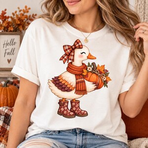 Peut inclure: Un t-shirt blanc avec une illustration de canard blanc portant un nœud à carreaux, une écharpe en tricot orange et des bottes à motifs floraux. Le canard tient dans ses ailes une citrouille orange ornée de feuilles d'automne et de fleurs.