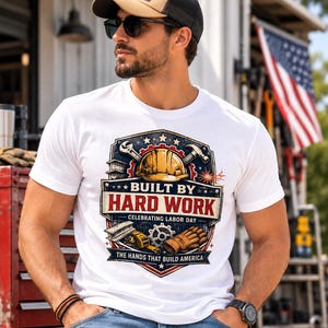 Peut inclure: Un t-shirt blanc avec un motif graphique comprenant un casque de chantier jaune, une clé, un marteau, un engrenage et un gant de travail. Le texte indique BUILT BY HARD WORK, CELEBRATING LABOR DAY et THE HANDS THAT BUILD AMERICA, avec des étoiles.