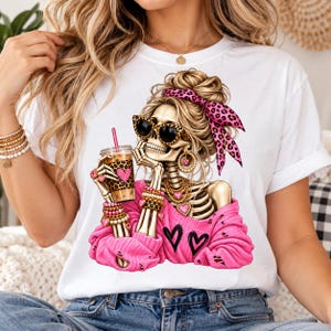 Può includere: Una t-shirt bianca con la grafica di uno scheletro con capelli biondi raccolti in uno chignon, una fascia rosa leopardata, occhiali da sole neri e un maglione rosa. Lo scheletro tiene in mano un bicchiere di caffè con una cannuccia rosa.