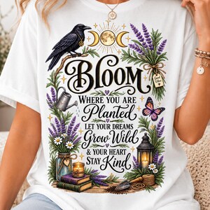 May include: A white t-shirt featuring a graphic print with the text Bloom Where You Are Planted Let Your Dreams Grow Wild & Your Heart Stay Kind. The design includes a black crow, a moon, lavender, a butterfly, a lantern, books, and a candle.