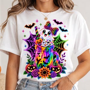 Peut inclure: Un t-shirt blanc avec un motif coloré représentant un fantôme portant un nœud à imprimé léopard, assis dans un bocal arc-en-ciel rempli de crème fouettée, de citrouilles et de chauves-souris, entouré de toiles d'araignée néon et de fleurs.