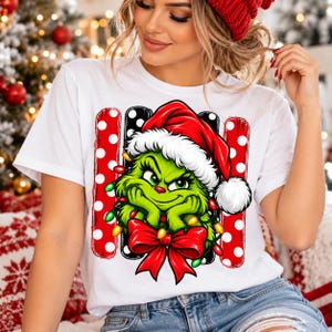 Könnte beinhalten: Ein weißes Kurzarm-T-Shirt mit einer Grafik einer grünen Figur, die eine rote Weihnachtsmütze trägt, verheddert in bunten Lichterketten, mit einer großen roten Schleife unten und einem Hintergrund aus roten und schwarzen vertikalen Streifen mit weißen Punkten.