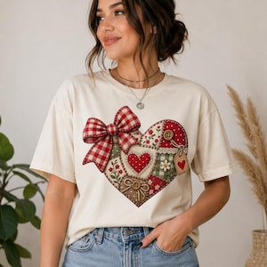 Peut inclure: Un t-shirt à manches courtes de couleur crème arborant un grand motif central en forme de cœur composé de tissus en patchwork. Le cœur présente un nœud à carreaux vichy rouge et blanc, des imprimés floraux, des pois et des boutons en bois.