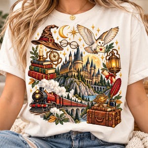 May include: A cream-colored t-shirt featuring a detailed graphic print of a castle, a steam train on a bridge, a stack of books, a witch hat, a flying owl with a letter, a lantern, a globe, a suitcase, and various stars and moon icons.