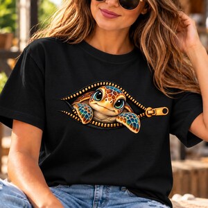 Puede incluir: Una camiseta negra de manga corta con un estampado gráfico de una pequeña tortuga marina asomándose detrás de una cremallera abierta. La tortuga tiene ojos grandes y un caparazón con patrones en tonos azules, naranjas y amarillos.