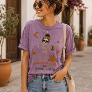 Peut inclure: Un t-shirt violet à manches courtes avec un motif de squelette portant un bandeau et des lunettes de soleil, tenant une tasse de café et faisant un signe de paix. Le design inclut une citrouille, des feuilles d'automne et le texte PUMPKIN SPICE AND good vibes.