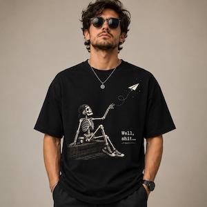 Peut inclure: Un t-shirt noir à manches courtes orné d'un motif représentant un squelette portant une casquette et des baskets, assis sur un rebord en briques avec une tasse de café. Le squelette tend la main vers un avion en papier. Le texte "Well, shit..." est inscrit.