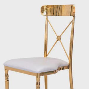 May include: A gold-colored metal chair with a white seat. The chair has an X-shaped back design with a decorative center and a curved top. The legs are also gold-colored.