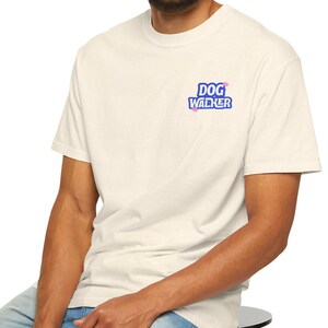 Puede incluir: Camiseta color crema con el texto "DOG WALKER" en azul y rosa. La camiseta la lleva una persona sentada en un taburete negro, con vaqueros azules. El fondo es blanco.