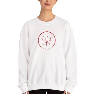 Puede incluir: Sudadera blanca con un diseño circular rojo que dice "OH CANADA". La sudadera es de cuello redondo y tiene mangas largas. El texto está escrito en un estilo manuscrito.