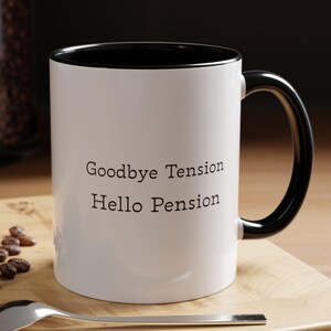 May include: White ceramic mug with a black interior and handle. The mug has the text "Goodbye Tension Hello Pension" in black. A spoon and a pastry are on a wooden surface.