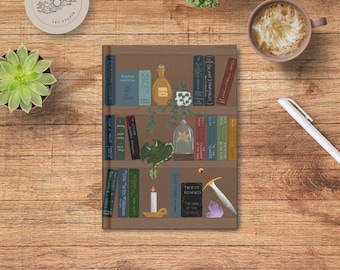 Può includere: Un taccuino con un design a scaffale con libri, una bottiglia e un drago. Sono raffigurati anche una spada, un cristallo e una candela. Il taccuino è su una superficie di legno, con una tazza di caffè, una penna e delle piante.