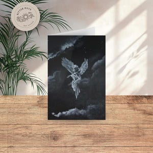 May include: A greeting card with a monochrome painting of an angel with outstretched wings against a dark, cloudy sky. The angel is in a flowing gown. The card is on a wooden surface.