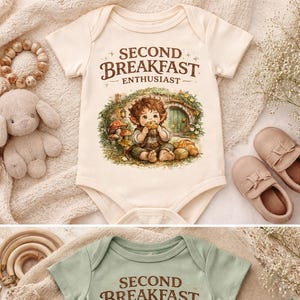 Op de afbeelding: Twee babyrompers, één crème en één saliegroen, elk met de tekst "SECOND BREAKFAST ENTHUSIAST". De afbeelding toont een kind dat een gebakje eet voor een deur die op een hobbitgat lijkt. De afbeelding heeft een grillige, sprookjesachtige uitstraling.