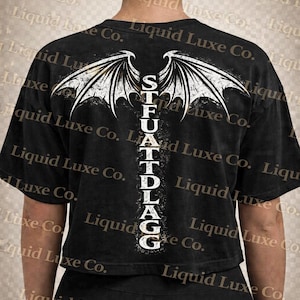 May include: Black crop top with a white graphic of stylized wings and the text "STFUATTDLAGG" in a vertical column. The shirt is made of a soft material and has short sleeves. The overall theme is edgy and alternative.