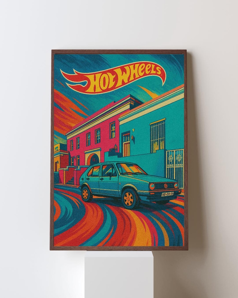May include: A framed print featuring a vintage teal car with orange wheels, parked on a street with colorful buildings. The Hot Wheels logo is displayed at the top. The artwork has a retro, stylized design with vibrant colors.