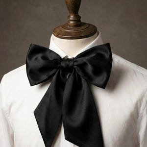 May include: A black satin bow tie, large in size, is displayed on a white collared shirt. The bow tie is the central element, offering a classic and refined appearance. The bow tie's design is symmetrical, with elongated, pointed tips.