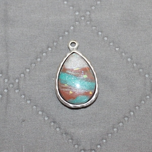 May include: A teardrop-shaped pendant with a silver-colored bezel. The pendant's interior features a mix of colors, including silver, teal, and reddish-brown, with a glittery effect. The pendant has a small loop at the top for a chain.