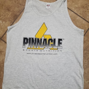 May include: A light gray tank top with the text "PINNACLE SPORTS NUTRITION" in black and yellow. The tank top has a large logo with a yellow and black design. The website address www.pinnaclesportsnutrition.com is printed below the text.