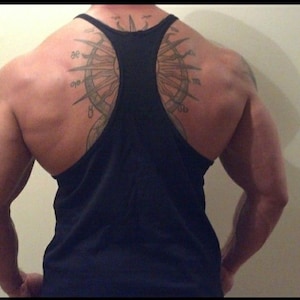 May include: A black tank top with a racerback design. The back features a large tattoo of a compass rose. The image shows the back of a person with muscular arms and shoulders.