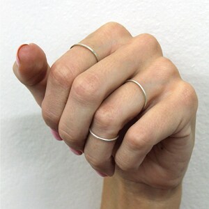 May include: A hand wearing three thin, silver rings. The rings are simple bands, with one on the index finger and another on the middle finger. The rings are delicate and minimalist in design. The background is a plain white.