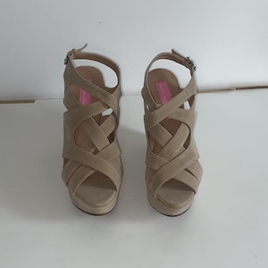 May include: A pair of beige platform sandals featuring a crisscross strap design across the vamp, an open toe, and an adjustable ankle strap with a metal buckle. The interior footbed includes a bright pink label with white text.