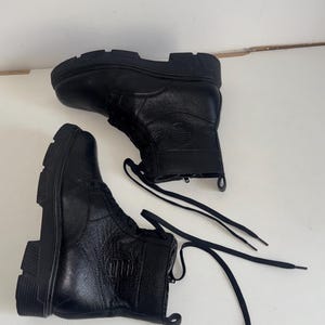 May include: A pair of black ankle boots featuring a textured leather upper, thick lugged rubber soles, and black laces. Each boot displays a circular embossed logo on the side, a pull tab at the heel, and a side zipper closure for easy access.