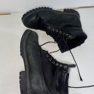 May include: A pair of black leather ankle boots featuring a lace-up front with silver-toned metal eyelets, a padded collar, and a thick, lugged rubber sole. The side of the boot displays a debossed circular logo, and the interior is lined with black fur.