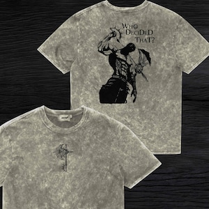 May include: A grey acid-wash t-shirt featuring a small black graphic of a battle axe on the front chest. The back of the shirt displays a large black illustration of a muscular figure holding a helmet with the text Who Decided That?