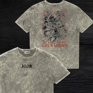 May include: Two grey t-shirts with a stone wash effect. One shirt features the word "JOJO" in black on the front. The other shirt has a graphic of characters and the text "WORLD 88" and "STAR DUST CRUSADERS" in red.