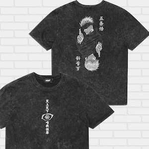 May include: Two dark grey t-shirts with Japanese text and anime-style illustrations. One shirt features a character with a blindfold, the other has an eye graphic. Both shirts have a crew neck and short sleeves.