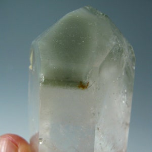 Puede incluir: Un cristal de cuarzo transparente con un tono verde en la parte superior. El cristal es puntiagudo en la parte superior y tiene una base plana.