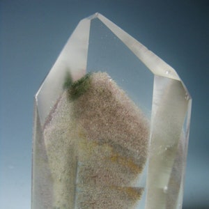 Puede incluir: Un cristal de cuarzo transparente con inclusiones de minerales marrones, verdes y amarillos. El cristal tiene forma de pirámide y una superficie lisa y pulida.