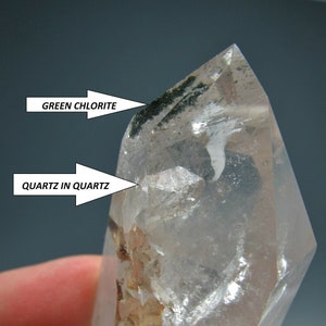 Puede incluir: Un cristal de cuarzo transparente con inclusiones de clorita verde y un cristal de cuarzo más pequeño en su interior. El texto "QUARTZ IN QUARTZ" y "GREEN CHLORITE" está escrito en la imagen.
