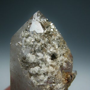 Puede incluir: Un cristal de cuarzo transparente con una superficie rugosa y texturizada. El cristal está cubierto de pequeños cristales blancos.