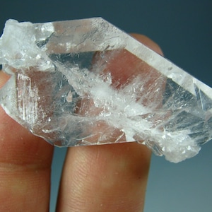 Puede incluir: Un cristal de cuarzo transparente con doble terminación. El cristal mide aproximadamente 5 cm de largo y tiene un brillo vítreo.