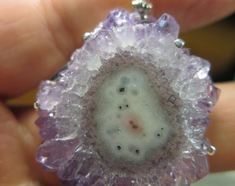 May include: A purple amethyst crystal pendant with a silver bail. The crystal has a natural, druzy finish and a white center.