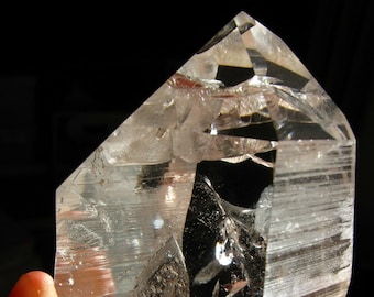May include: A clear quartz crystal with a unique, faceted shape. The crystal is held in a hand, showcasing its intricate details and natural beauty.