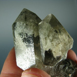 Puede incluir: Dos cristales de cuarzo transparentes con un color gris ahumado y un aspecto nublado y lechoso. Los cristales están agrupados y tienen una textura rugosa y natural.