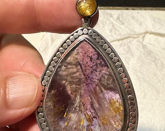 Op de afbeelding: Een zilveren hanger met een traanvormige, iriserende edelsteen met gouden vlekken. De hanger heeft een parelrand en een kleinere, ronde, gouden edelsteen aan de bovenkant.