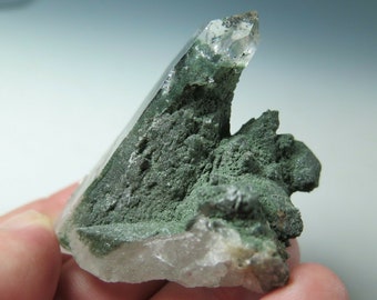 Może przedstawiać: Przezroczysty kryształ kwarcu z zielonym mineralnym nalotem. Kryształ jest ostry i szpiczasty, a nalot jest szorstki i fakturowany.