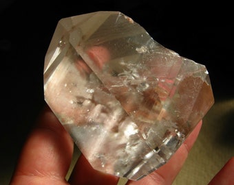 May include: A large, clear quartz crystal with a double termination. The crystal is translucent and has a slight pink hue. It is a natural crystal and has a rough, unpolished surface.