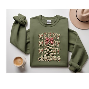 Puede incluir: Una sudadera de cuello redondo color verde oliva con un estampado gráfico de un árbol de Navidad hecho de cuerda trenzada, rematado con un lazo a cuadros rojo y blanco. El texto MERRY MERRY MERRY y christmas está impreso en una fuente floral.