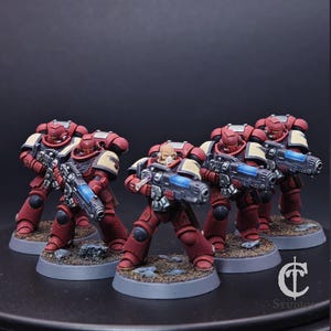 May include: Five miniature figures in red armour with beige shoulder pads and blue energy weapons. The figures stand on grey bases with a textured, brown surface. The figures are posed as if in action.