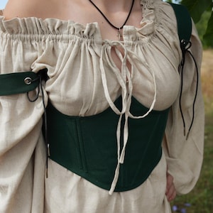 May include: A beige off-the-shoulder blouse with long sleeves and a drawstring neckline, paired with a dark green corset-style waist cincher. The sleeves feature dark green bands with metal eyelets and black cord ties on the upper arms.